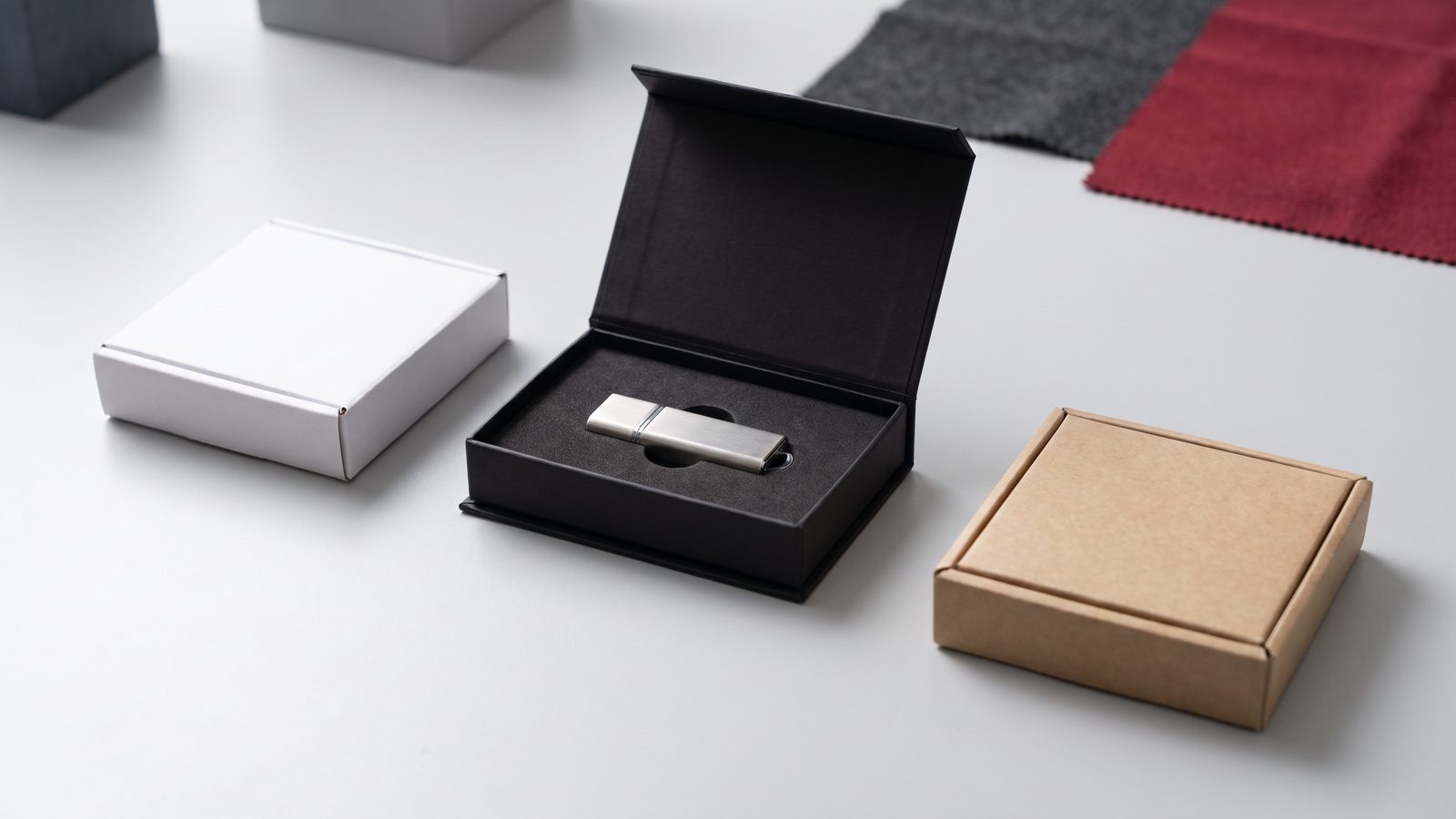 Ilustrační kovový USB disk v otevřené černé krabičce vedle bílé a kraftové krabičky